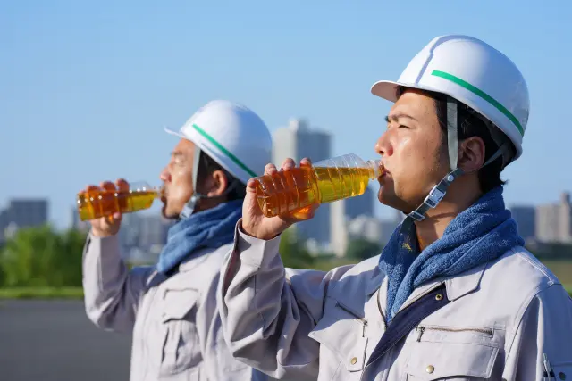 炎天下でペットボトルの麦茶を飲む男性工事作業員2名。